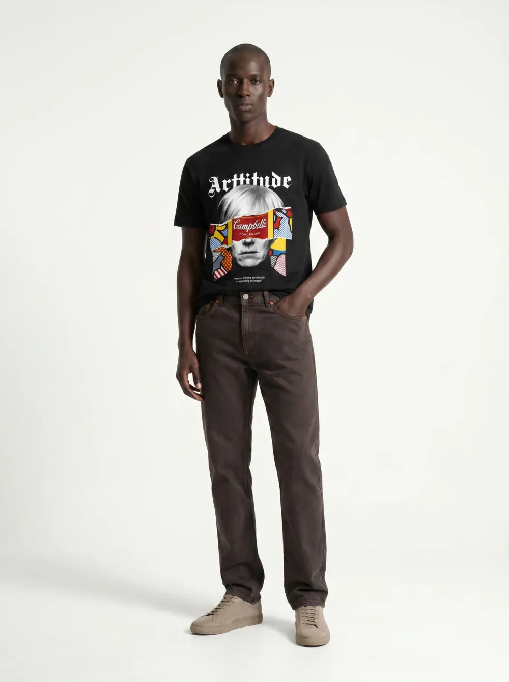 Black tee with a colour portrait print, brown trousers, tan sneakers, one hand in a pocket